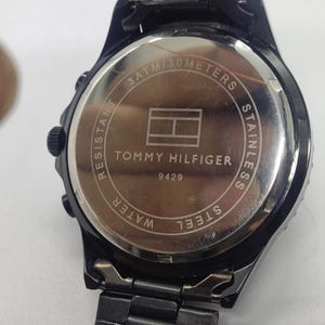 May include: Black Tommy Hilfiger watch with a stainless steel back. The watch face features the Tommy Hilfiger logo and the text "3 ATM/30 METERS". The watch band is black and made of metal.