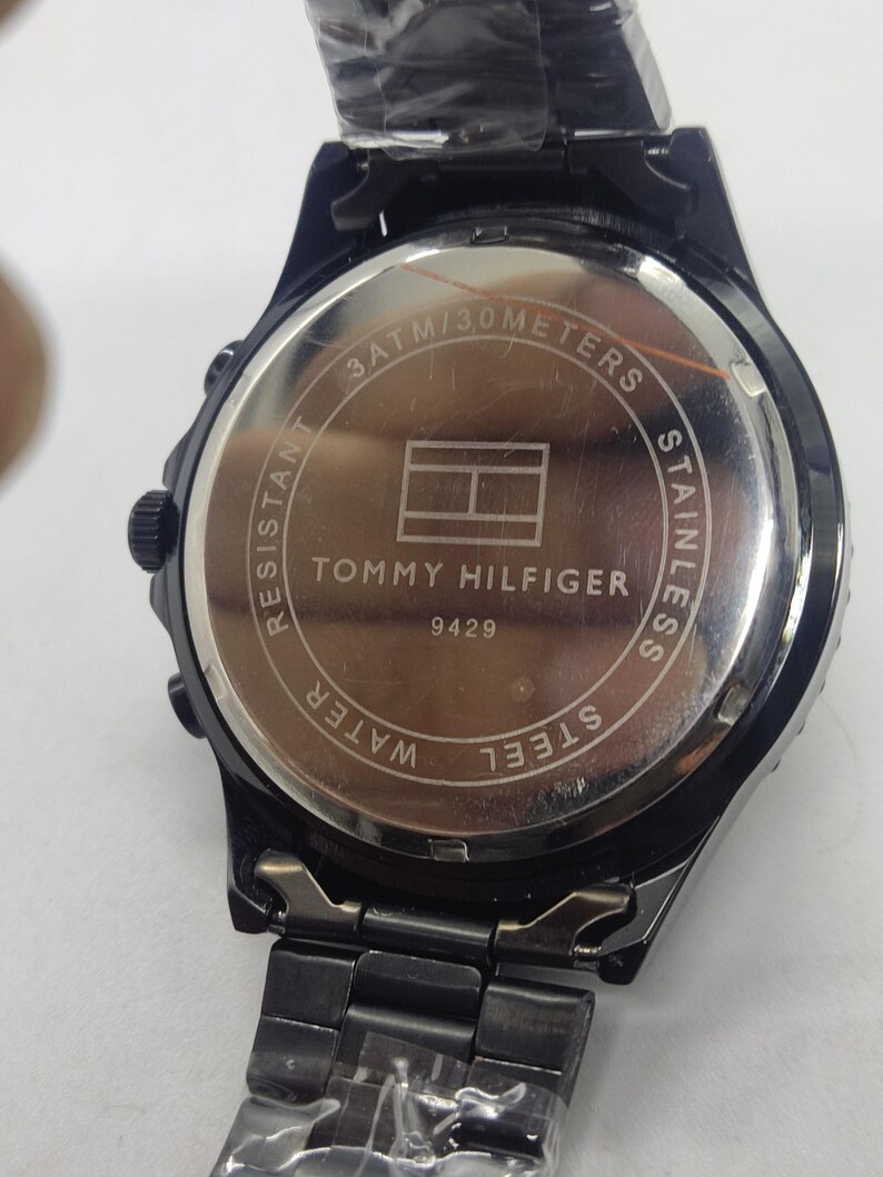 May include: Black Tommy Hilfiger watch with a stainless steel back. The watch face features the Tommy Hilfiger logo and the text "3 ATM/30 METERS". The watch band is black and made of metal.