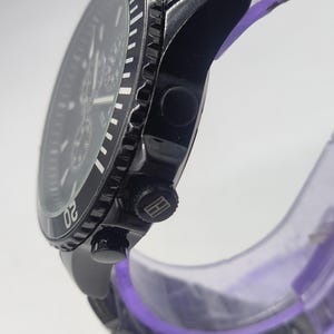 May include: A black wristwatch with a stainless steel band. The watch face is black with white markings and a rotating bezel. The watch has a chronograph function and a logo on the crown. The watch is displayed on a clear purple stand.