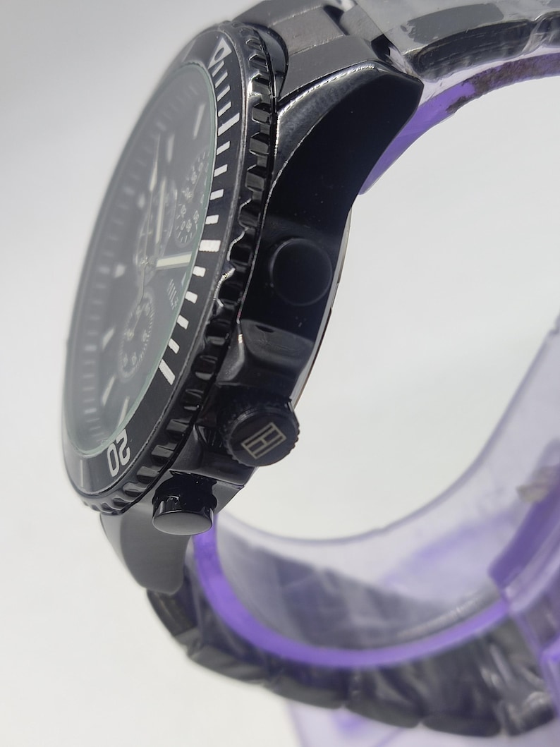May include: A black wristwatch with a stainless steel band. The watch face is black with white markings and a rotating bezel. The watch has a chronograph function and a logo on the crown. The watch is displayed on a clear purple stand.