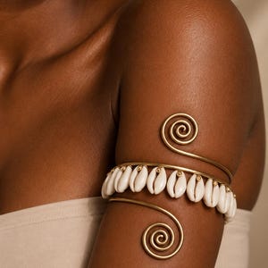 May include: A gold-toned metal arm cuff featuring a spiral design at both ends and a central row of white cowrie shells. The arm cuff is worn on the upper arm, and a matching cowrie shell earring is visible near the shoulder area.
