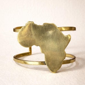 May include: A gold-toned metal cuff bracelet featuring a large, flat, brushed-finish cutout of the African continent centered on two parallel horizontal bands. The text LEGACY Design is printed in dark brown at the bottom of the image.