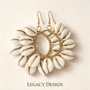 May include: A pair of handcrafted earrings featuring white cowrie shells arranged around a gold-toned wire frame. The earrings have gold-colored ear wires and the text "LEGACY DESIGN" at the bottom.
