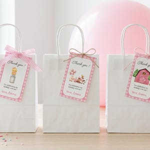 May include: Three white paper gift bags with handles, each adorned with a pink tag and a decorative bow or twine. The tags feature illustrations and the text "Thank you!" and "love, Emma". A pink balloon is in the background.