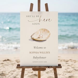 以下が含まれることがあります： 木製のイーゼルに立てられたクリーム色の看板。テキストは「She'll be here soon」と「Welcome to SOPHIA WILLIS'S Babyshower」です。看板には、真珠が入った開いた貝殻の水彩イラストが描かれています。日付は2026年7月25日土曜日です。