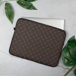 May include: A brown laptop sleeve with a repeating pattern of interlocking shapes and a bicycle-like design in a lighter brown color. The sleeve is open, revealing a white laptop inside. The sleeve has a black zipper.