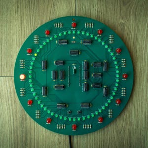 May include: A round, green circuit board with numerous black chips and red and green LED lights. The board is set against a wooden background. The lights are arranged in a circular pattern, suggesting a clock or timer.