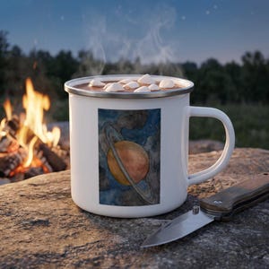 May include: A white enamel mug with a silver rim, filled with a hot beverage topped with white marshmallows. The mug features a rectangular graphic of a planet with rings set against a dark blue, starry, watercolor-style space background.
