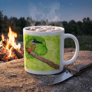 May include: A white ceramic mug filled with a hot chocolate beverage topped with white marshmallows. The mug features a watercolor-style illustration of a green bird perched on a brown branch against a vibrant green background.