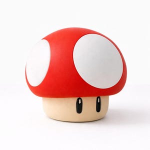 Può includere: Un oggetto decorativo a forma di fungo rosso con due grandi macchie bianche sul cappello. La base è beige con due occhi neri ovali. Ispirato alla serie di videogiochi Super Mario.