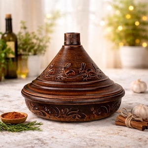 Puede incluir: Una olla de tagine de cerámica marrón con tapa cónica y tallas florales. La olla se encuentra sobre una superficie de color claro, con un pequeño cuenco de madera con especias y ramitas de romero cerca. Una botella de vino está en el fondo.