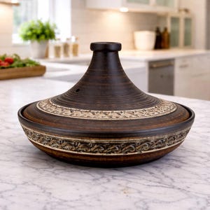 Può includere: Un tagine in ceramica marrone scuro con coperchio conico e fascia decorativa attorno alla base. La pentola è su un piano di lavoro in marmo in una cucina. Il tagine ha un design tradizionale, adatto per cucinare e servire piatti marocchini.