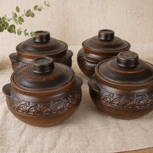 May include: Four brown ceramic cooking pots with lids. The pots have a round shape with handles and decorative floral patterns. The lids have a small knob on top. The pots are arranged on a beige surface.