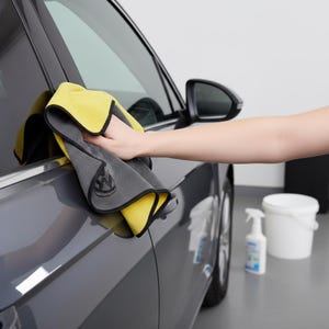 Puede incluir: Un paño de limpieza de coche de microfibra amarillo y gris se utiliza para limpiar una puerta de coche gris. El paño tiene un ribete negro y un logotipo. Un pulverizador y un cubo blanco están en el fondo.