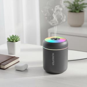 Peut inclure: Un humidificateur gris foncé avec un dessus aux couleurs de l'arc-en-ciel et le mot "HUMIDIFIER" imprimé sur le côté. L'appareil émet de la vapeur blanche. D'autres éléments sur la table comprennent une petite plante succulente, un carnet et des écouteurs blancs.