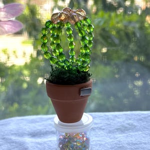 May include: A small beaded cactus sculpture in a terracotta pot. The cactus is made of green beads, with amber-coloured beads at the top. The pot sits on a small jar filled with iridescent beads.