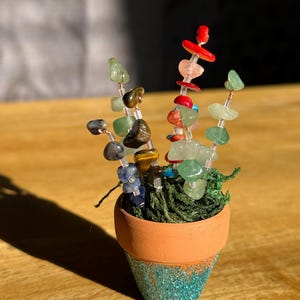 May include: A miniature terracotta pot with a teal glitter base, filled with green moss and three wire stems adorned with assorted polished gemstone chips in shades of blue, brown, red, and light green, arranged in a vertical, plant-like structure.