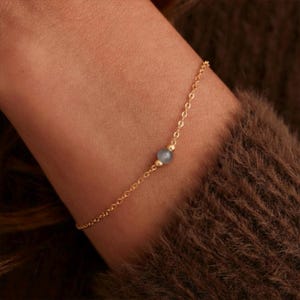 Può includere: Un delicato braccialetto a catena color oro con una piccola perla rotonda azzurra al centro. Il braccialetto è indossato su un polso, su uno sfondo di tessuto marrone strutturato.
