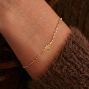 Peut inclure: Un bracelet doré délicat avec un petit cœur. Le cœur a un motif texturé. Le bracelet est porté sur un poignet, et l'arrière-plan est un flou brun.