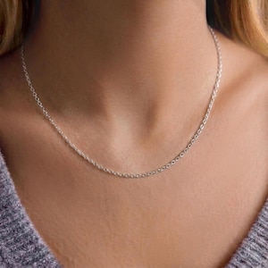Puede incluir: Un delicado collar de cadena de plata. El collar es un diseño de cadena simple y delgada, que descansa sobre el pecho de una persona. La cadena es de color plateado brillante y parece estar hecha de metal.