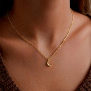 Pode incluir: Um colar dourado com um pequeno pingente de lua crescente. A corrente é fina e o pingente é pequeno, criando um design minimalista e elegante. O colar é usado em volta do pescoço.