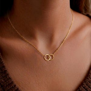 Peut inclure: Un collier doré délicat avec deux cercles entrelacés. Le pendentif est suspendu à une fine chaîne, créant un design simple mais élégant. Le collier est présenté sur un fond marron.