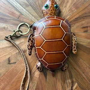 May include: A brown leather turtle-shaped keychain with a gold-colored keyring and strap. The turtle has a patterned shell design with a light brown outline and green eyes. The keychain is on a wooden surface.