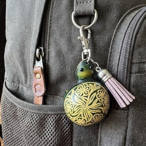 May include: A decorative keychain with a green turtle-shaped top and a round, yellow and green patterned body. The keychain has a silver clasp and a pink tassel, attached to a gray backpack.