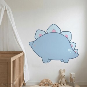 Könnte beinhalten: Ein großer, hellblauer Dinosaurier-Wandtattoo mit einem freundlichen Gesichtsausdruck. Der Cartoon-Stegosaurus hat hellblaue Platten mit rosa Akzenten. Der Aufkleber wird an einer weißen Wand in einem Kinderzimmer gezeigt.