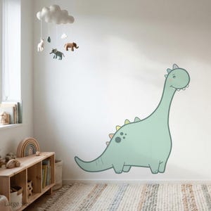 Könnte beinhalten: Ein großer, hellgrüner Cartoon-Dinosaurier-Wandtattoo mit langem Hals und freundlichem Lächeln. Der Dinosaurier hat bunte Akzente und befindet sich an einer weißen Wand in einem Kinderzimmer. Ein Mobile mit Tierfiguren hängt darüber.