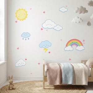 Könnte beinhalten: Eine Kinderzimmerwand mit bunten Cartoon-Wetterelementen dekoriert, darunter eine lächelnde Sonne, Wolken, ein Regenbogen und Regentropfen. Rosa Herzen sind über die Wand verstreut. Ein Holzbett mit Pastelldecken und ein gewebter Teppich vervollständigen die Szene.