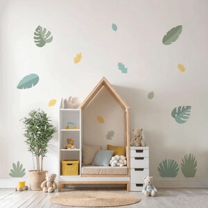 Könnte beinhalten: Ein Kinderzimmer mit einem hausförmigen Bettrahmen aus Holz, weißen Regalen und einer kleinen weißen Kommode. Der Raum ist mit Blättern in Grün-, Gelb- und Blautönen dekoriert. Eine Topfpflanze, Teddybären und eine gelbe Aufbewahrungsbox ergänzen die Einrichtung.