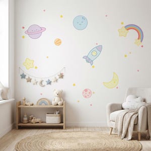 Könnte beinhalten: Ein Kinderzimmer mit einer weißen Wand, die mit Cartoon-Planeten, Sternen und einem Regenbogen geschmückt ist. Ein Holzregal beherbergt Spielzeug, und ein beiger Sessel mit einem Wolkenkissen und einer Decke steht auf einem runden Jute-Teppich. Der Raum ist in sanften Pastellfarben gehalten.