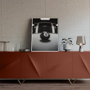 May include: Black and white framed art print featuring an abstract image of an eight ball. The artwork is displayed on a red-brown cabinet with gold legs, complemented by a small potted plant and a modern lamp.