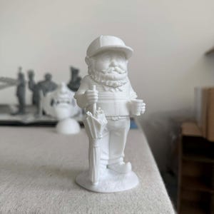 Könnte beinhalten: Eine weiße Figur eines bärtigen Mannes mit Mütze, der einen Regenschirm und eine Tasse hält. Die Figur steht auf einem kleinen, runden Sockel. Weitere weiße Figuren sind im Hintergrund zu sehen.