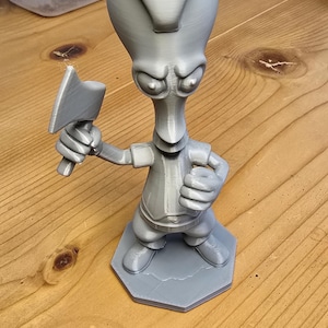 May include: A gray 3D-printed figurine of a cartoon alien character holding a small axe. The figure is standing on a gray hexagonal base. The figurine is on a wooden surface.
