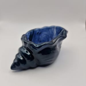 May include: A glossy, dark navy blue ceramic bowl shaped like a conch shell. The exterior features a deep, reflective dark blue glaze, while the interior reveals a lighter, marbled blue finish with a smooth, undulating rim and a spiral point.