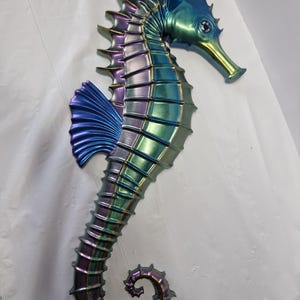 May include: A wall-mounted seahorse sculpture featuring a metallic, iridescent finish that shifts between shades of deep purple, vibrant blue, and emerald green. The figure displays a segmented body, a curved tail, and a prominent dorsal fin.