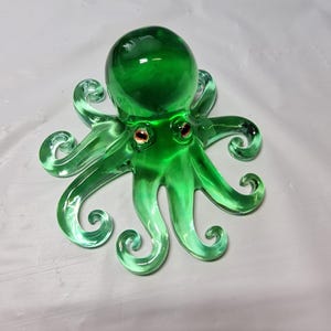 May include: A vivid green glass octopus ornament featuring eight curled tentacles and two red eyes. The octopus has a rounded head. It is displayed on a white surface. This decorative piece is a unique home decor item.