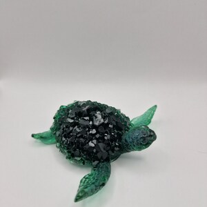May include: A sea turtle figurine featuring a translucent green glass head and flippers with a textured, scaly surface. The shell is covered in a cluster of dark, faceted black crystals that create a sharp contrast against the smooth green glass.
