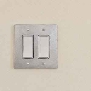 May include: A double light switch with two white rectangular rockers set into a square, textured silver-colored metal wall plate. The plate is secured to the wall with four small, visible silver screws positioned at the top and bottom corners.