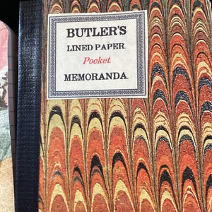 May include: A pocket notebook with a marbled cover featuring orange, yellow, and black arched patterns. A white rectangular label on the front reads BUTLER'S LINED PAPER Pocket MEMORANDA in black and red text, bordered by a decorative frame.