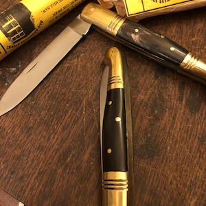 May include: A collection of vintage knives with brass accents and dark wooden handles. One knife is open, revealing a silver blade. The packaging reads "KNIFE" and "AT THE SIGN OF THE MILL SAW". The knives are arranged on a wooden surface.