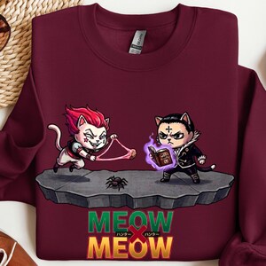 May include: Burgundy sweatshirt with a graphic featuring two cat-like characters, a spider, and the text "MEOW MEOW" in green and yellow. One character holds a string, the other a book.