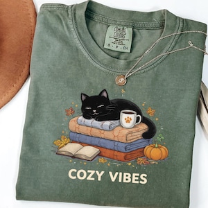 Puede incluir: Camiseta verde Comfort Colors con un gráfico de un gato negro durmiendo sobre mantas, una taza de café y una calabaza. El texto "COZY VIBES" está impreso en la parte inferior. Un collar dorado está sobre la camiseta.