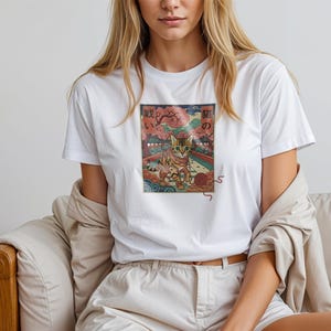 May include: White t-shirt featuring a colorful graphic of a cat in a Japanese-inspired landscape. The design includes a cat, cherry blossoms, and Japanese text. The shirt is made of a soft material.