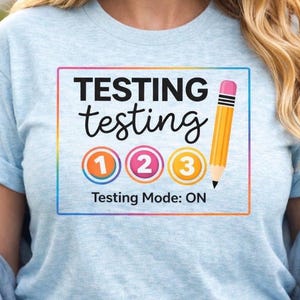 Puede incluir: Camiseta azul claro con un gráfico colorido que dice "TESTING testing 1 2 3 Testing Mode: ON" y una ilustración de lápiz. La camiseta está hecha de un material suave.