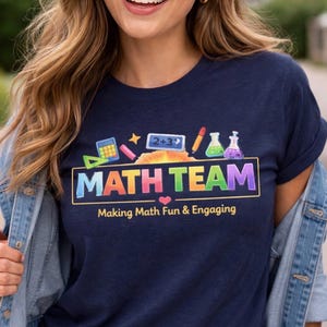 Könnte beinhalten: Marineblaues T-Shirt mit der Aufschrift "MATH TEAM" in bunten Buchstaben und mathematischen Grafiken. Der Text "Making Math Fun & Engaging" steht darunter. Das T-Shirt wird getragen.