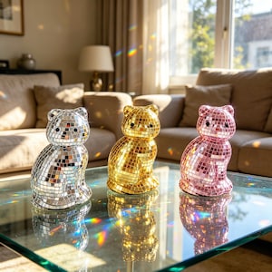 May include: Three cat-shaped decorative objects. One is silver, one is gold, and one is pink. Each cat is covered in small, reflective squares, creating a disco ball effect. They sit on a glass table, reflecting light.