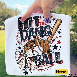 Peut inclure: T-shirt blanc avec un motif sur le thème du baseball. Le design comprend une batte, un gant, une balle de baseball et le texte "Hit the Dang Ball" en noir et rouge. Un nœud et des étoiles complètent le design. Le t-shirt a le texte "Wear Your Identity".